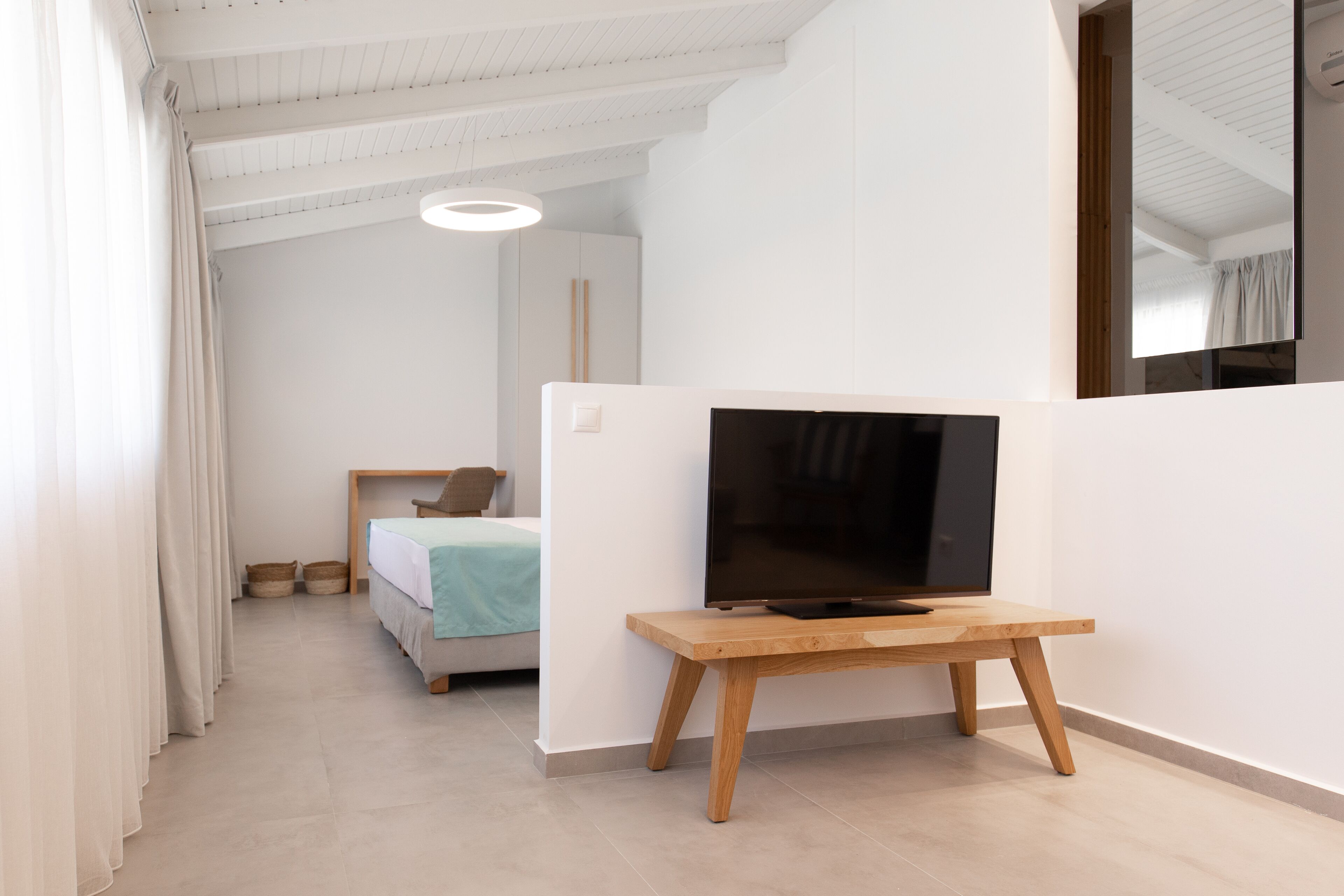 A modern, minimalist room with a bed, a wooden table with a flat-screen TV, and a large window.
