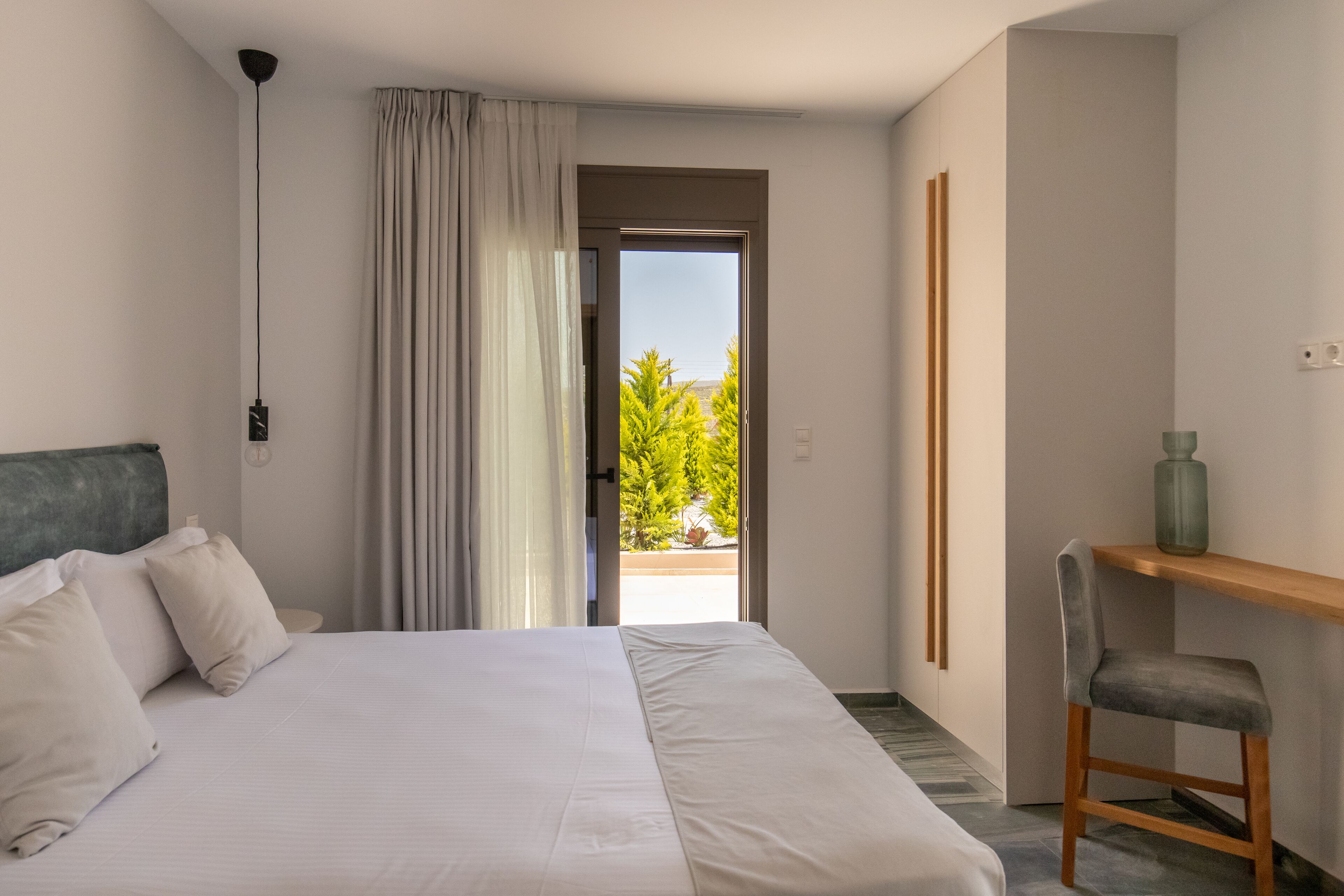 Ein Schlafzimmer mit Bett, Stuhl, Schreibtisch und Blick auf Grünanlage durch eine Tür.