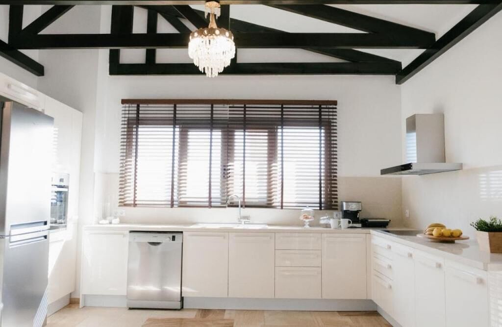A modern kitchen with white cabinetry, a built-in dishwasher, a large window with blinds, and a chandelier.