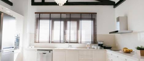 A modern kitchen with white cabinetry, a built-in dishwasher, a large window with blinds, and a chandelier.