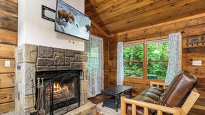 Ein gemütliches Blockhaus-Interieur mit Kamin aus Naturstein, einem Ledersessel und einem großen Fenster mit Blick auf den Wald.