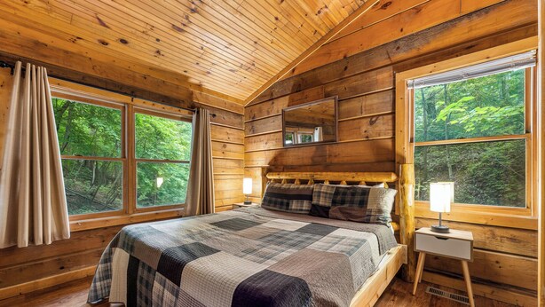 Ein Schlafzimmer einer Holzhütte mit einem großen Bett, zwei Fenstern, einem Nachttisch und einem Spiegel.