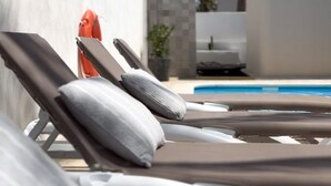 Ein Poolbereich mit Liegestühlen und einem kleinen Tisch, eine weiße Wand und eine Palme im Hintergrund.