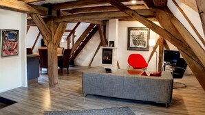 Een zolderkamer met zichtbare houten balken, een open haard, een grijze bank en rode stoelen.