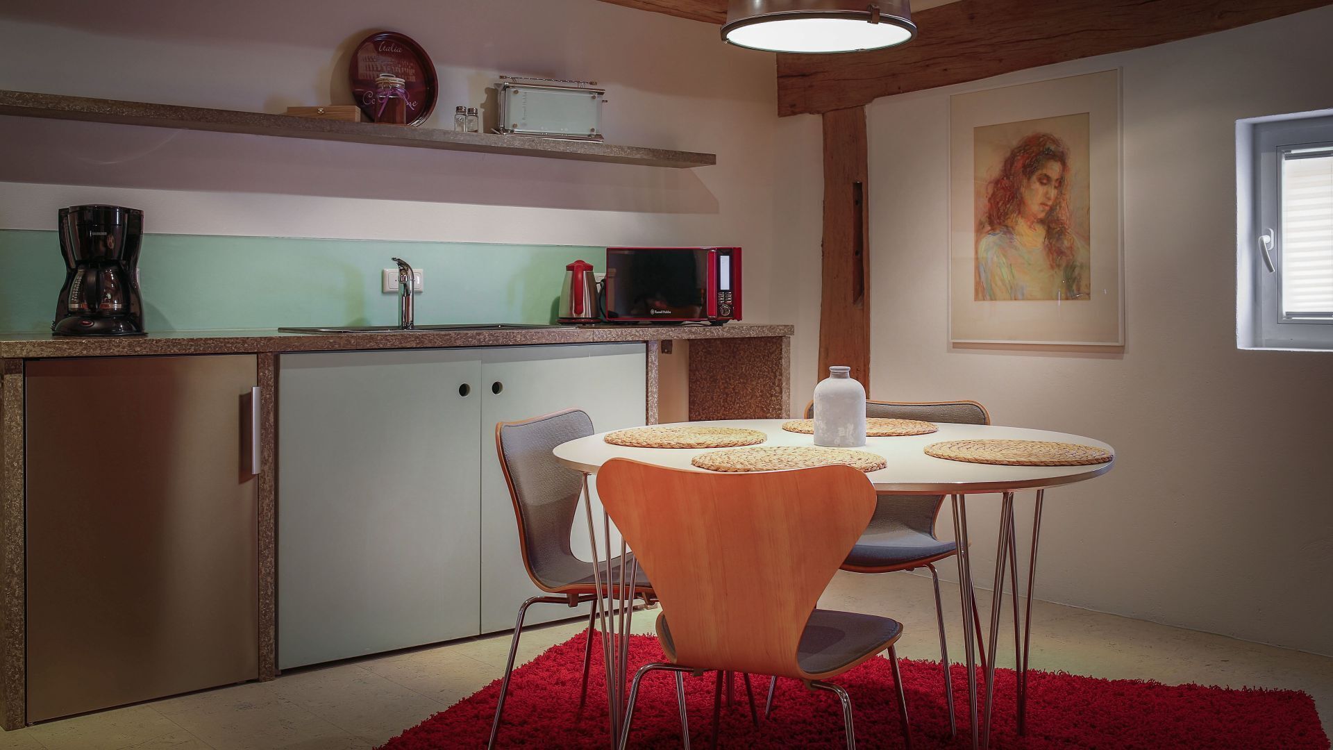 Een compacte keuken met een eethoek. Er is een magnetron, koffiezetapparaat en een kleine koelkast aanwezig. De eettafel is gedekt met geweven placemats en een witte vaas.