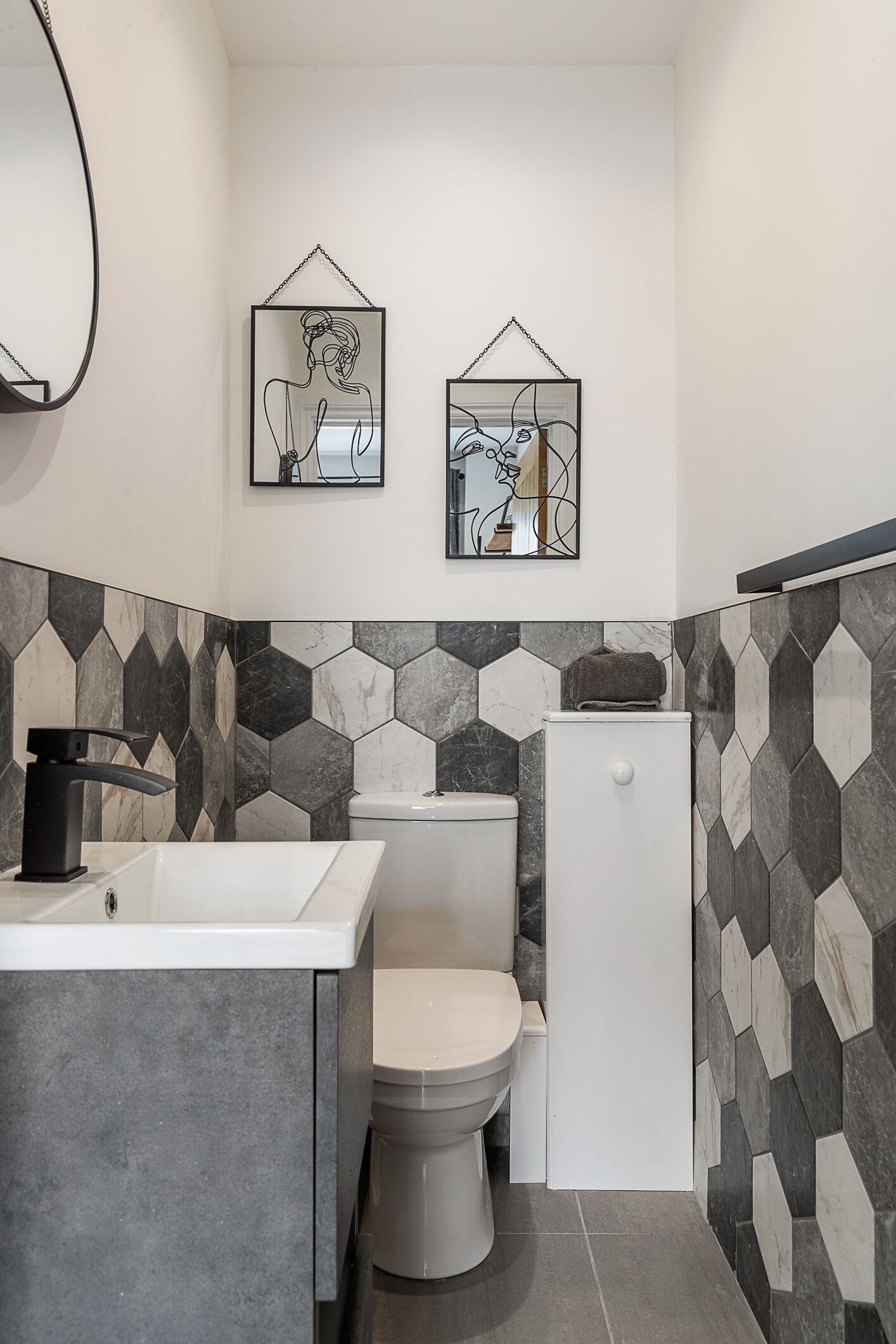 A modern bathroom with a white sink, toilet, and shower. There are two framed artworks on the wall.