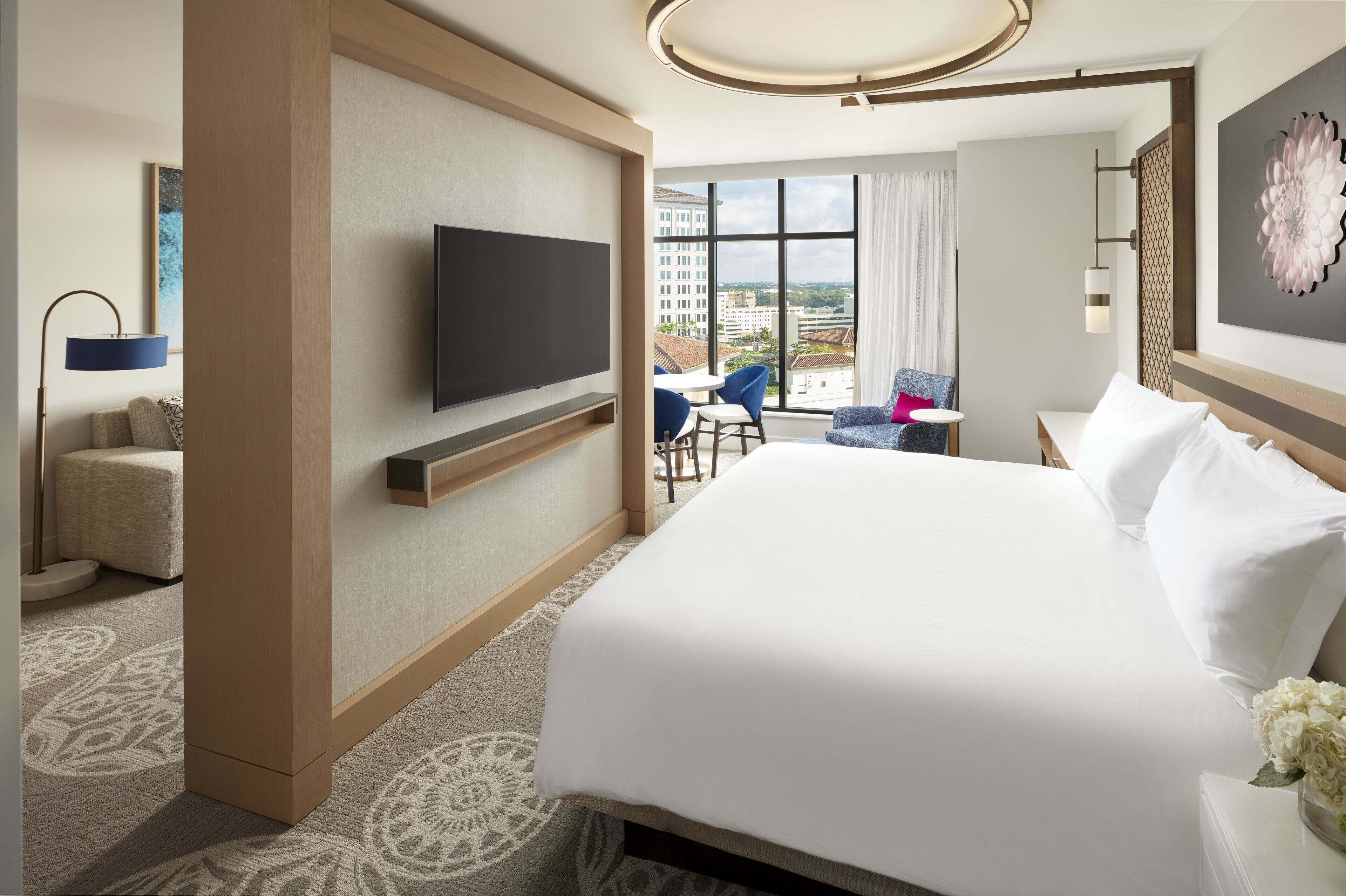 A hotel room with a large bed, a flat-screen TV mounted on the wall, a seating area with a table and chairs, and a view of the city through large windows.