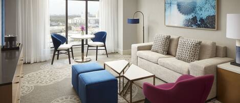 A modern hotel room with a large window, a sofa, a coffee table, and a dining area with chairs.