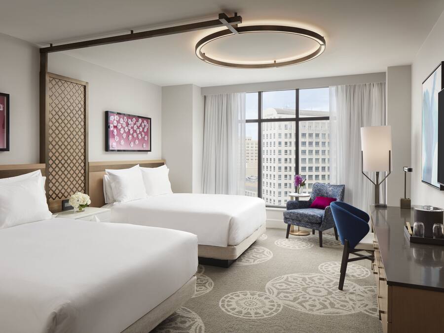 A hotel room with two beds, a desk, a chair, a TV, and a large window with a city view.