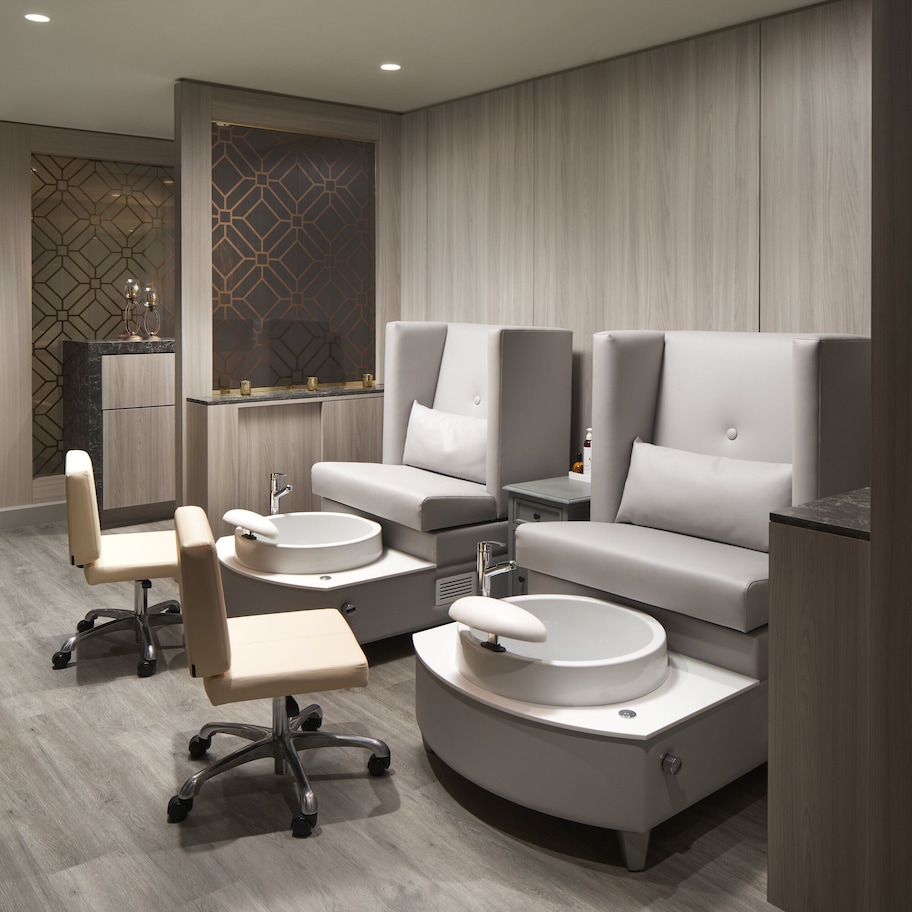 A modern spa room with a large mirror, two massage chairs, a small table, and a vanity with a sink.