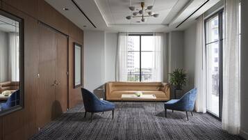 Ein modernes Hotelzimmer mit einem großen Fenster, einem braunen Sofa, zwei blauen Sesseln und einem gemusterten Teppich.