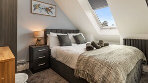 A bedroom with a sloped ceiling, a bed with a grey headboard, a bedside lamp, a framed artwork, and a window with a view of the sky. - Sycamore Cottage (Tenby)
