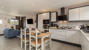A modern kitchen with white cabinets, a wooden dining table, and a built-in oven. - Sycamore Cottage (Tenby)
