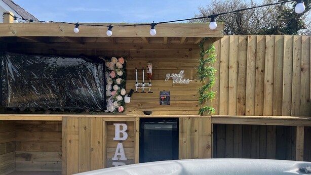 A wooden outdoor hot tub area with a wooden deck, a sign reading "good vibes," and a decorative wall with flowers and candles. - Sycamore Cottage (Tenby)