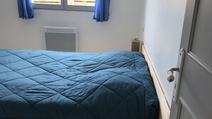 A single-bed room with a blue bedspread, a window with blue curtains, a radiator, and a small picture on the wall. - Cosy semi-detached house, terrace 800m from the swimming pool (Lanton)