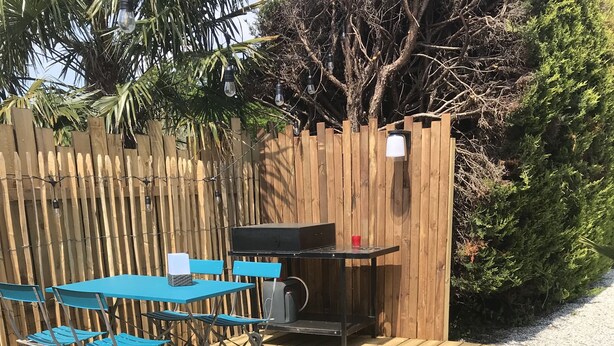 A wooden deck with a blue table and chairs, a grill, and a small table with a red cup. - Cosy semi-detached house, terrace 800m from the swimming pool (Lanton)