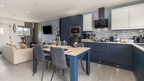 A modern kitchen with a dining area, featuring a built-in oven, microwave, and a flat-screen TV. - Birch Cottage (Tenby)