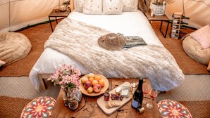 Un alojamiento tipo tienda de campaña acogedor con una cama, almohadas, una mesita de noche con una botella de vino, una bandeja de frutas y una tabla de quesos.