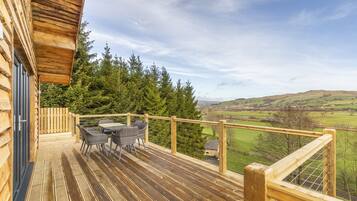 A wooden deck with a table and chairs, overlooking a green landscape with trees and hills.