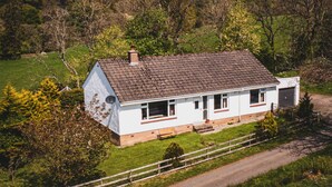 A white two-story house with a grey roof, surrounded by greenery and a hill in the background. - Keppoch Cottage (Crieff)