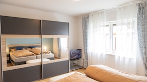 A modern hotel room with a bed, a TV, a mirror, and a window with curtains. - Beautiful newly renovated vacation apartment in a quiet location (Wald-Michelbach)