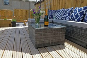 Une terrasse en bois avec une table à plateau de verre, une bouteille de champagne et un vase de fleurs.