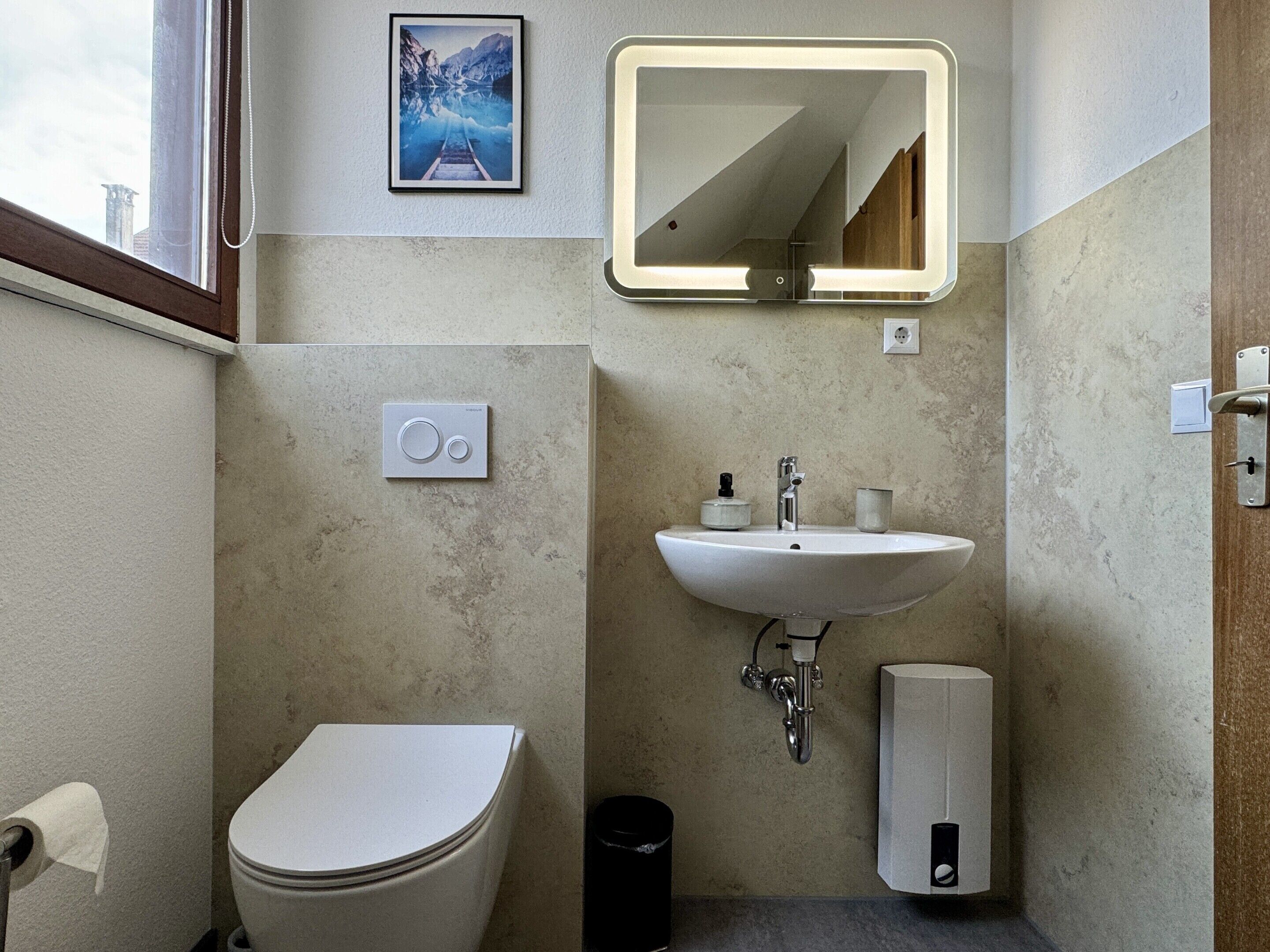 A bathroom with a toilet, sink, mirror, and a picture on the wall.