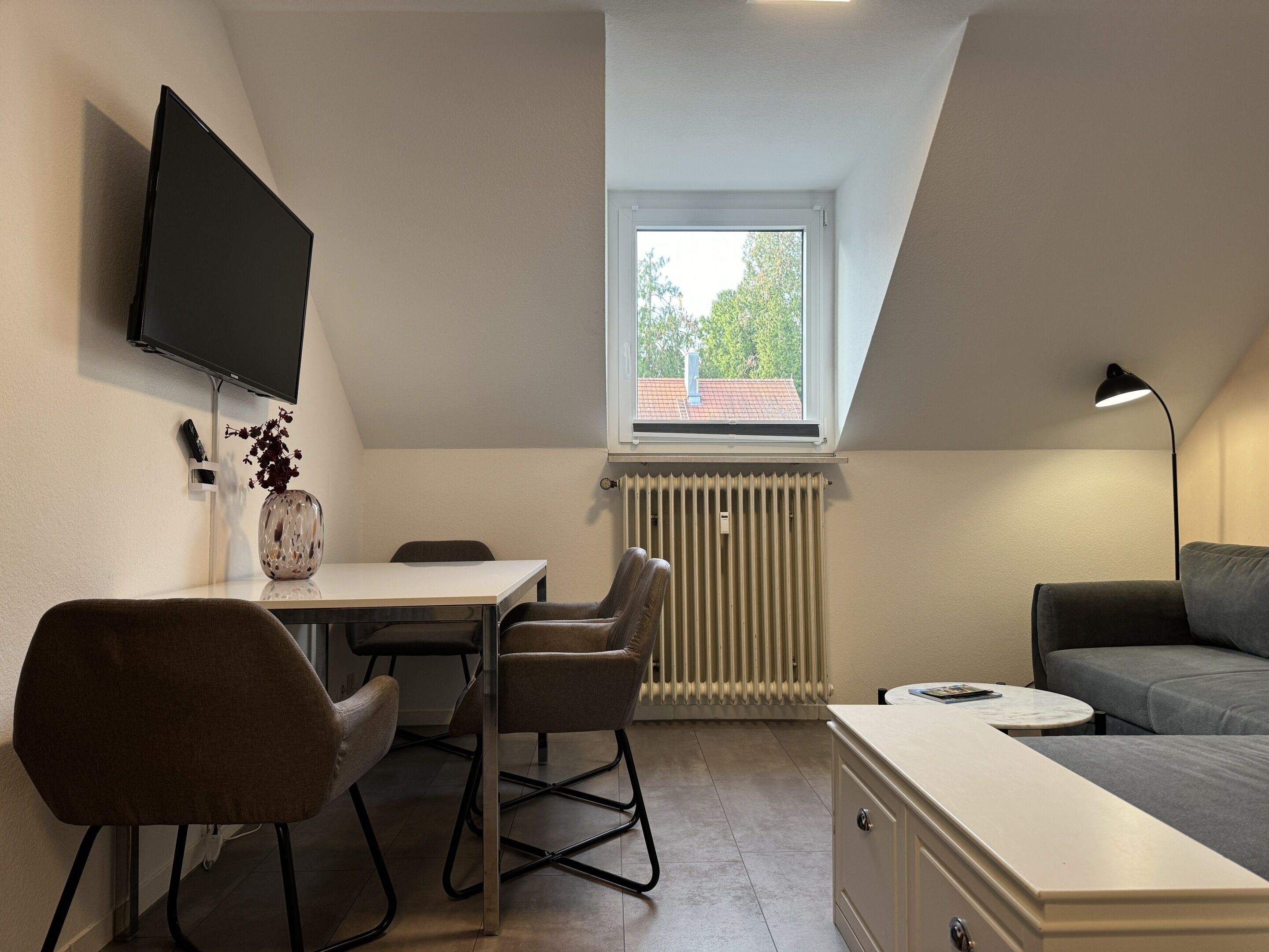 A compact living space with a dining area, a sofa, a TV mounted on the wall, and a window with a view of trees.