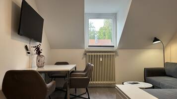 A compact living space with a dining area, a sofa, a TV mounted on the wall, and a window with a view of trees.