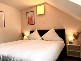Cama bem arrumada com lençóis brancos em quarto com teto inclinado, quadro na parede e mesa de cabeceira com abajur e um vaso.