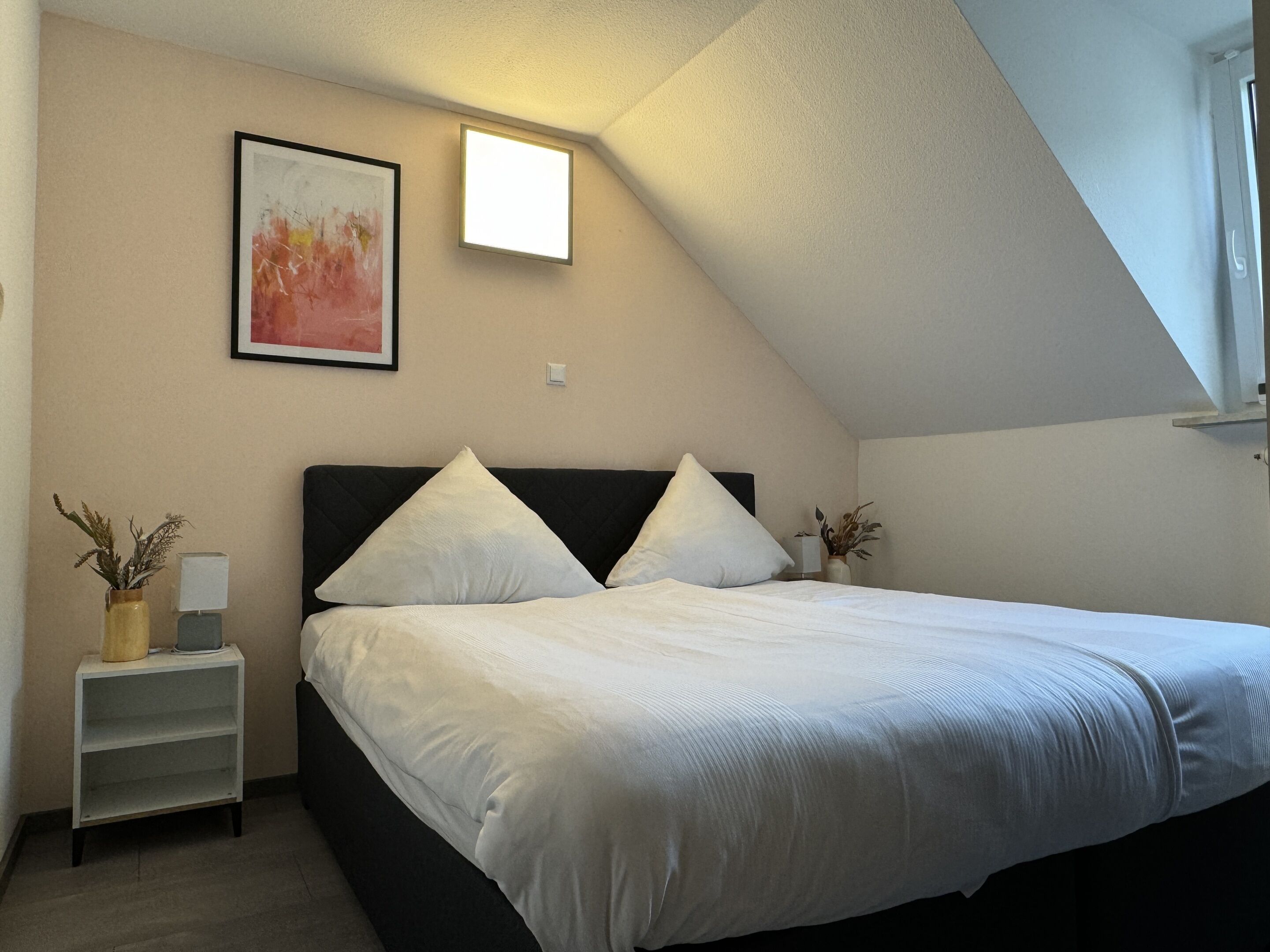 A bedroom with a bed, a nightstand, a wall-mounted light, and a framed picture.
