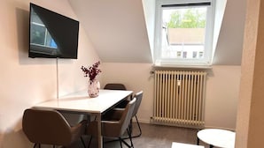 A room with a dining table, chairs, a TV mounted on the wall, a window with a view of greenery, and a radiator.