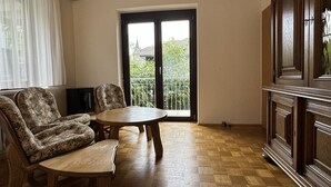 A room with a wooden floor, a round table, two floral-patterned chairs, a cabinet, and a sliding glass door leading to a balcony with greenery outside. - Ferienhaus Möhrle 4 (Ueberlingen)