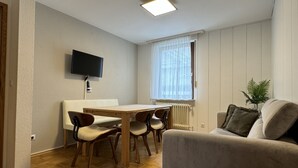 A living room with a sofa, a dining table with chairs, a TV mounted on the wall, a window with sheer curtains, and a radiator. - Ferienhaus Möhrle 4 (Ueberlingen)