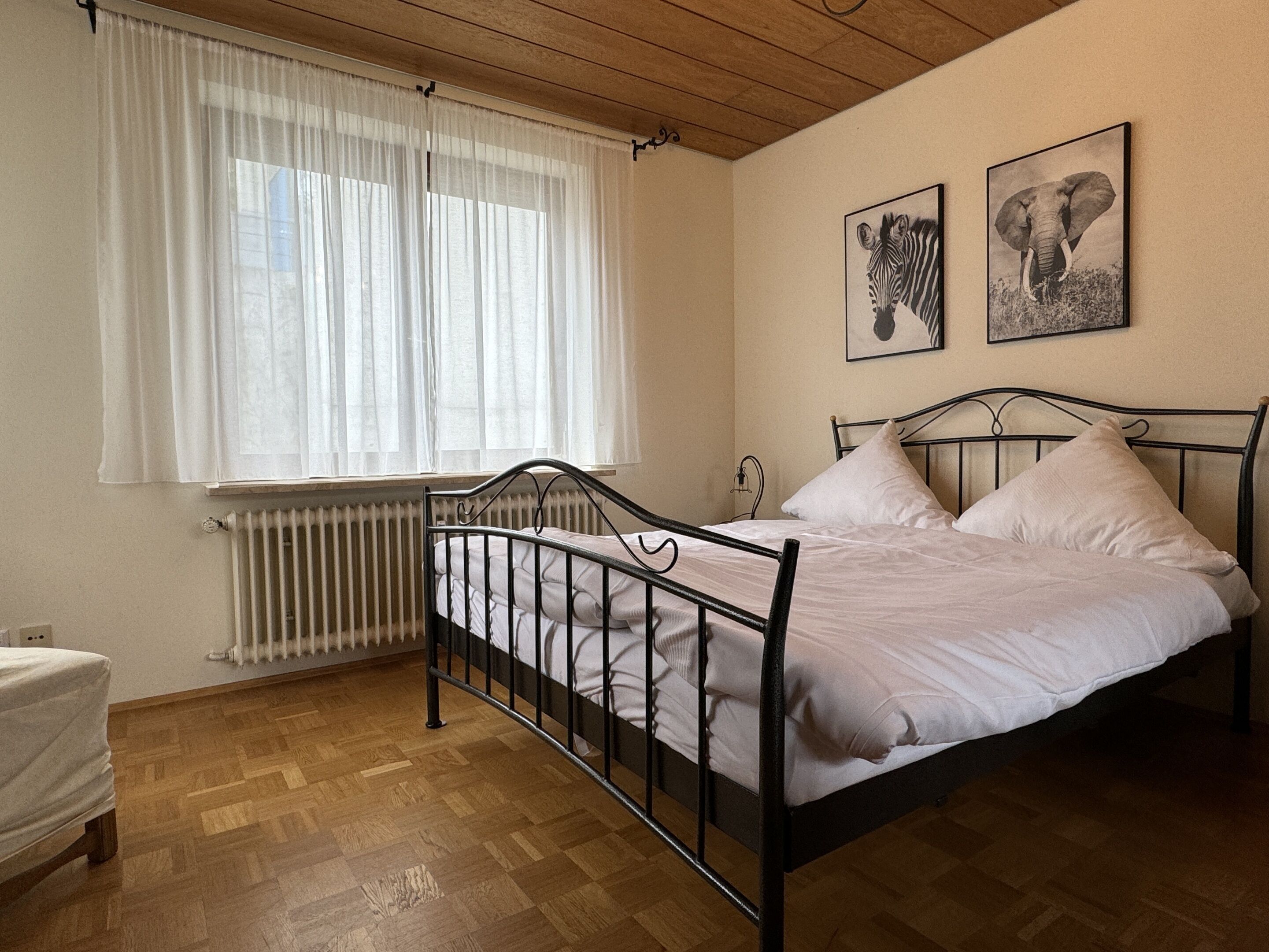 A bedroom with a metal bed frame, white bedding, a radiator, a window with sheer curtains, and two framed animal pictures on the wall.