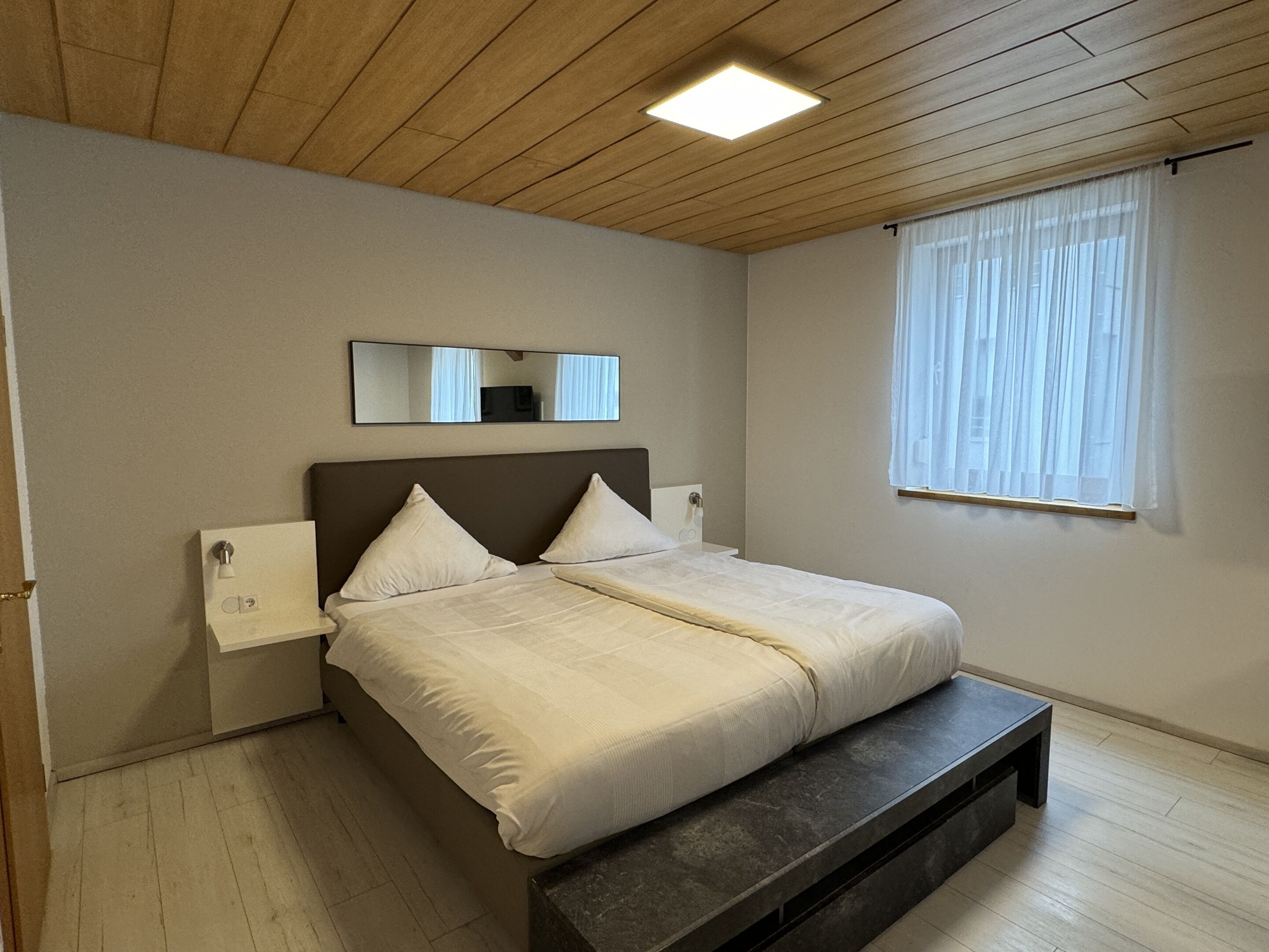 A bedroom with a bed, a bench, a nightstand, a mirror, and a window with curtains.