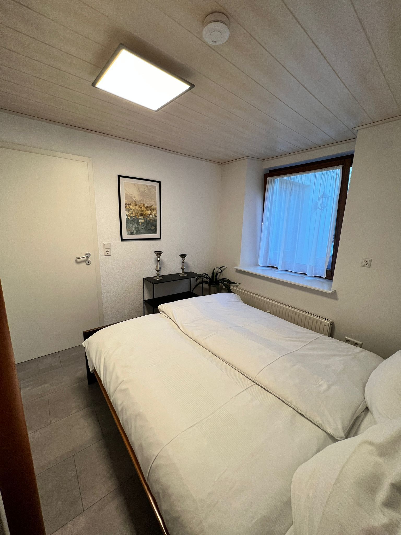 A bedroom with a bed, a bedside table with a lamp, a window with curtains, and a ceiling light.