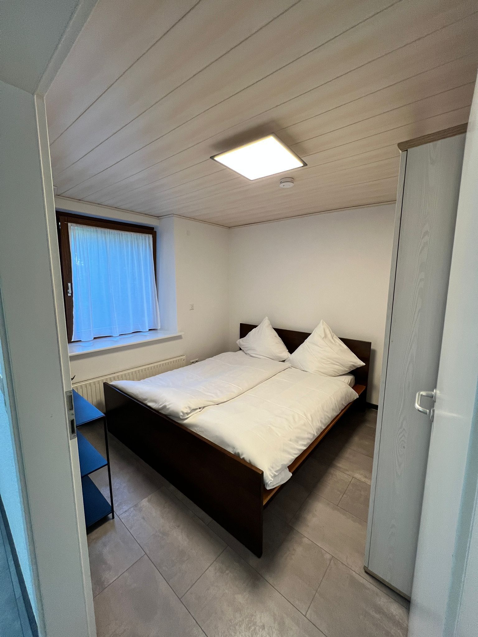 Un dormitorio con cama, ventana con cortinas, radiador y armario.