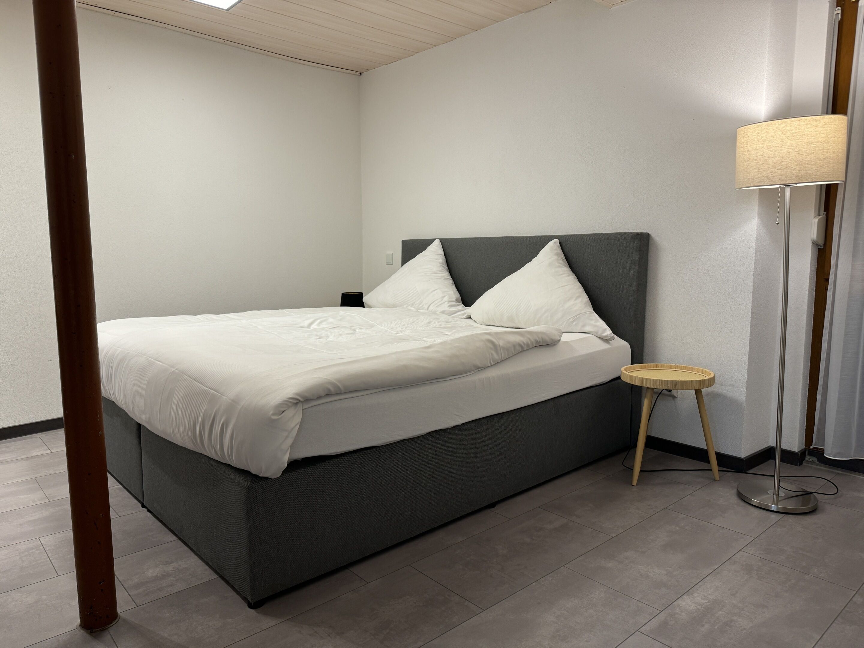 Een moderne slaapkamer met een grijs bed, witte lakens, een rond houten nachtkastje en een vloerlamp.