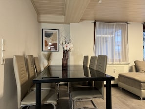 Uma sala de estar moderna com uma mesa de jantar de vidro rodeada por cadeiras de couro, um sofá bege, uma pintura abstrata emoldurada e um vaso com flores brancas.