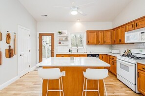A modern kitchen with wooden cabinets, a white countertop, a central island with bar stools, a microwave, a stove, and a ceiling fan.