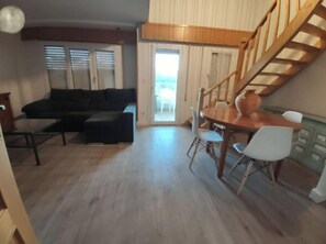 Un salon avec un canapé de couleur sombre, une table à manger en bois et quatre chaises, ainsi qu’un escalier menant à l’étage supérieur.