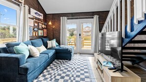 A modern living room with a blue sofa, a checkered rug, a TV on a wooden stand, and a staircase with blue railings. - Beach Cottage · Hot Tub · Rooftop · Movie Room (Kill Devil Hills)
