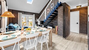 A dining area with a wooden table set for four, white chairs, a blue sofa, and a staircase with a blue railing. - Beach Cottage · Hot Tub · Rooftop · Movie Room (Kill Devil Hills)
