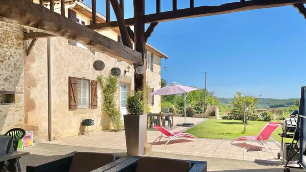 Un patio al aire libre con mobiliario de mimbre, una mesa y una silla. Hay dos sombrillas, una planta en maceta y una vista de un paisaje verde al fondo.