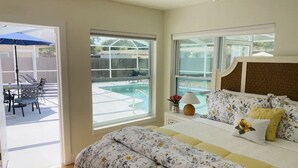 A bedroom with a large bed, a nightstand, a lamp, and a view of a swimming pool through the window. - Private Pool and Close to Clearwater beach! Perfect for any family! (Clearwater)