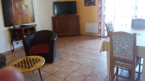 Een woonkamer met een televisie op een houten kast, een zwarte fauteuil, een kleine ronde tafel en een eettafel met stoelen.