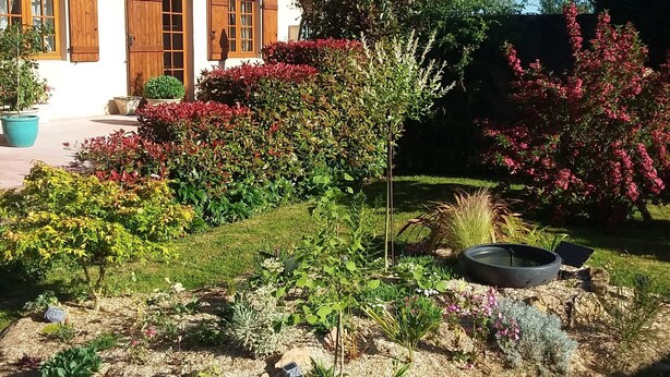 Een huis met een pannendak, houten luiken en een verzorgde tuin met diverse planten en een kleine boom.
