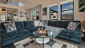 A modern living room with a sectional sofa, a glass coffee table, and large windows offering a view of the landscape. - Near Jordanelle & Deer Valley with Mountain & Lake Views, Hot Tub, Game Room & More! (Heber City)