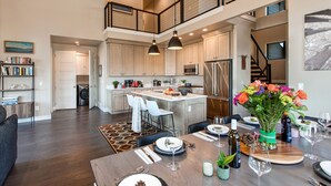 A spacious kitchen with a central island, a dining area with a wooden table set for a meal, and a staircase leading to an upper level. - Near Jordanelle & Deer Valley with Mountain & Lake Views, Hot Tub, Game Room & More! (Heber City)